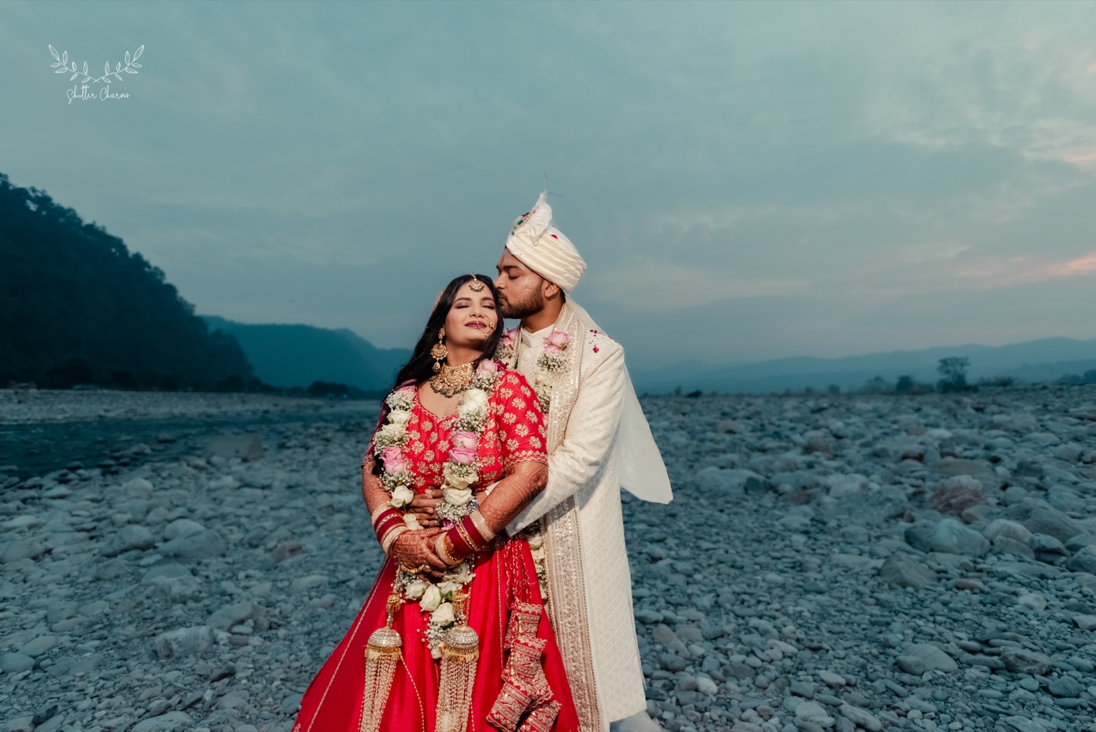 Groom kissing bride's forehead on a scenic riverbed at dusk