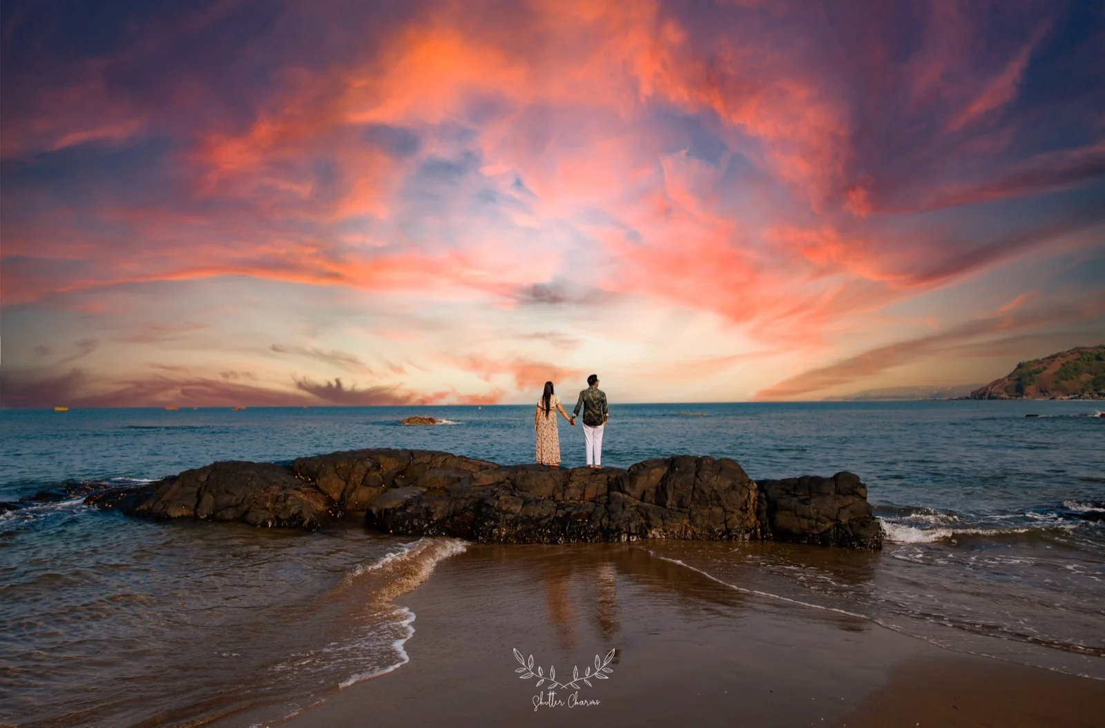 Couple holding hands on beach rocks beneath a dramatic sunset sky